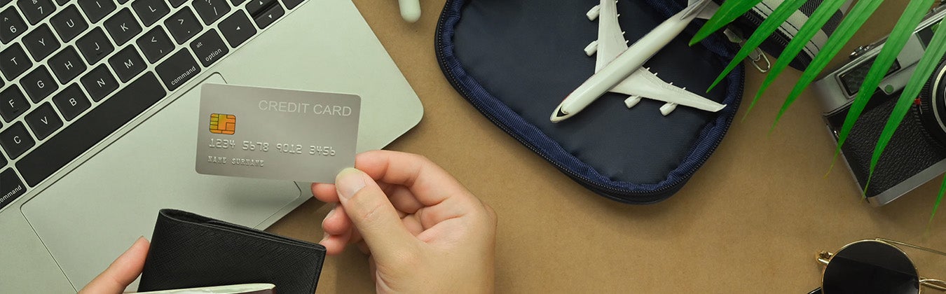 Person holding a credit card in front of an open laptop, with a camera, sunglasses and other travel props.